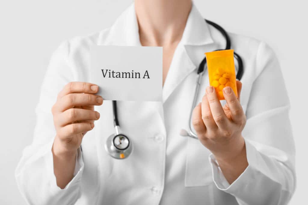 Female doctor holding paper with text VITAMIN A and pills on light background