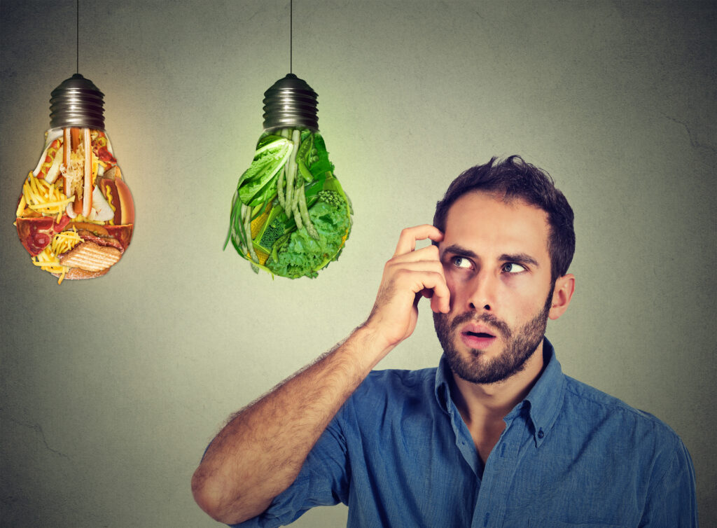 Puzzled man thinking looking up at junk food and green vegetables shaped as light bulbs making decision isolated on gray background. Diet choice right nutrition healthy lifestyle wellness concept.